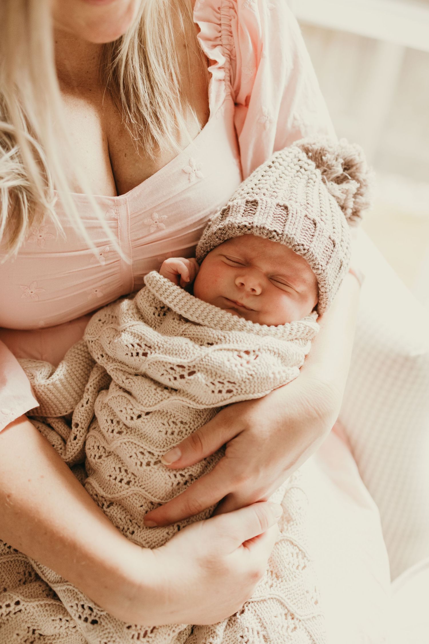 Sleeping newborn wrapped in knitted beige blanket and matching hat being cradled in gentle embrace.