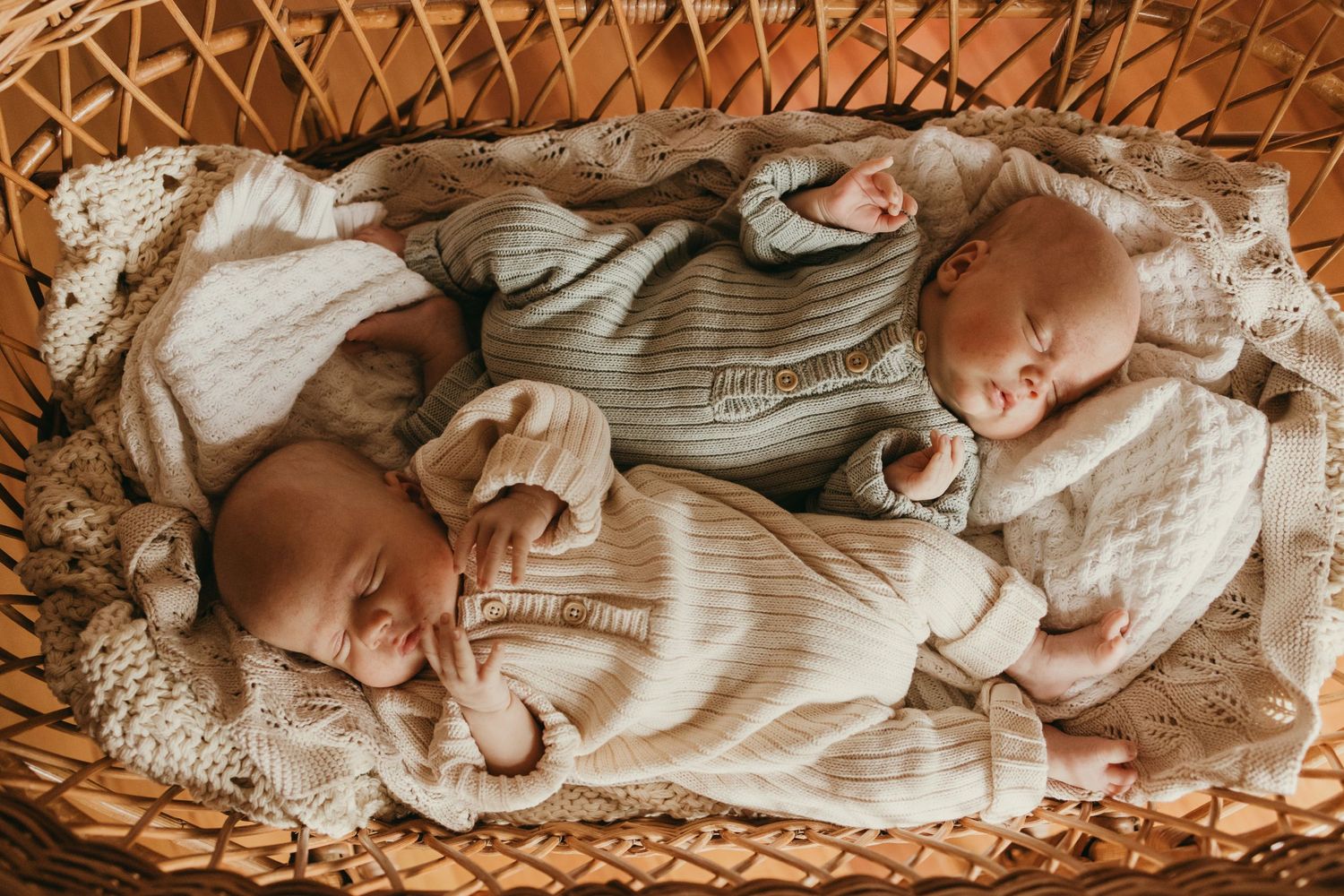 Twin babies in coordinated grey sweaters sleeping peacefully in a wicker bassinet.