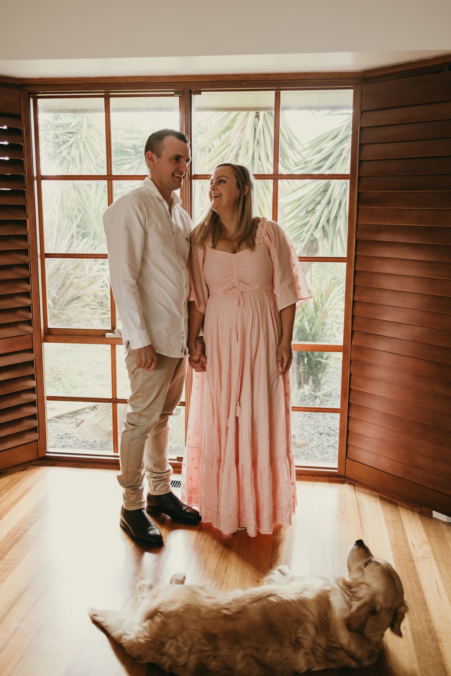 Couple standing by window with dog lying on hardwood floor.