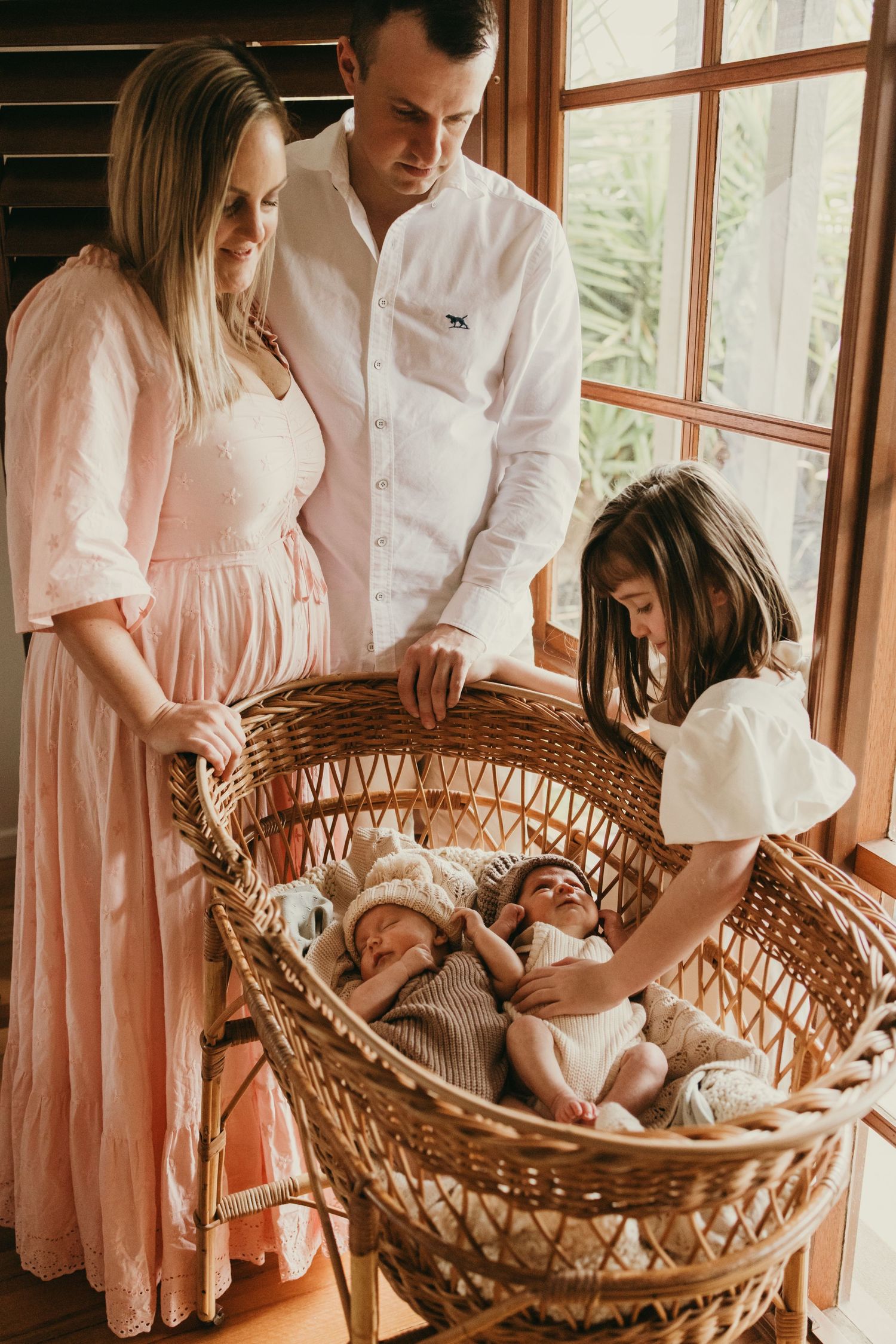 Parents and child looking down at newborn twins resting in woven bassinet.