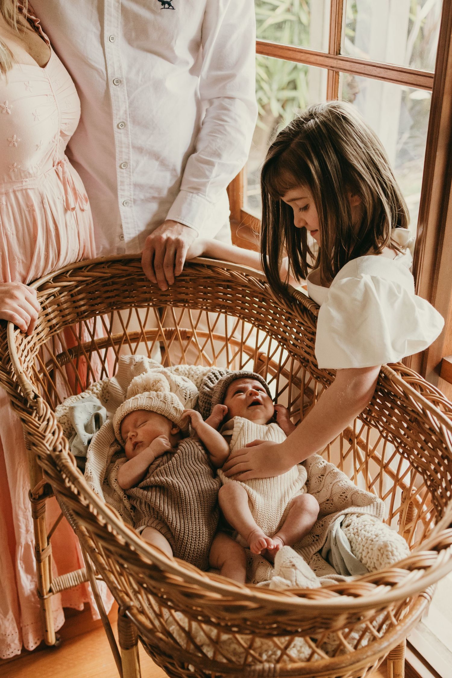 Family gathering around wicker bassinet with sleeping twins in knitted outfits.