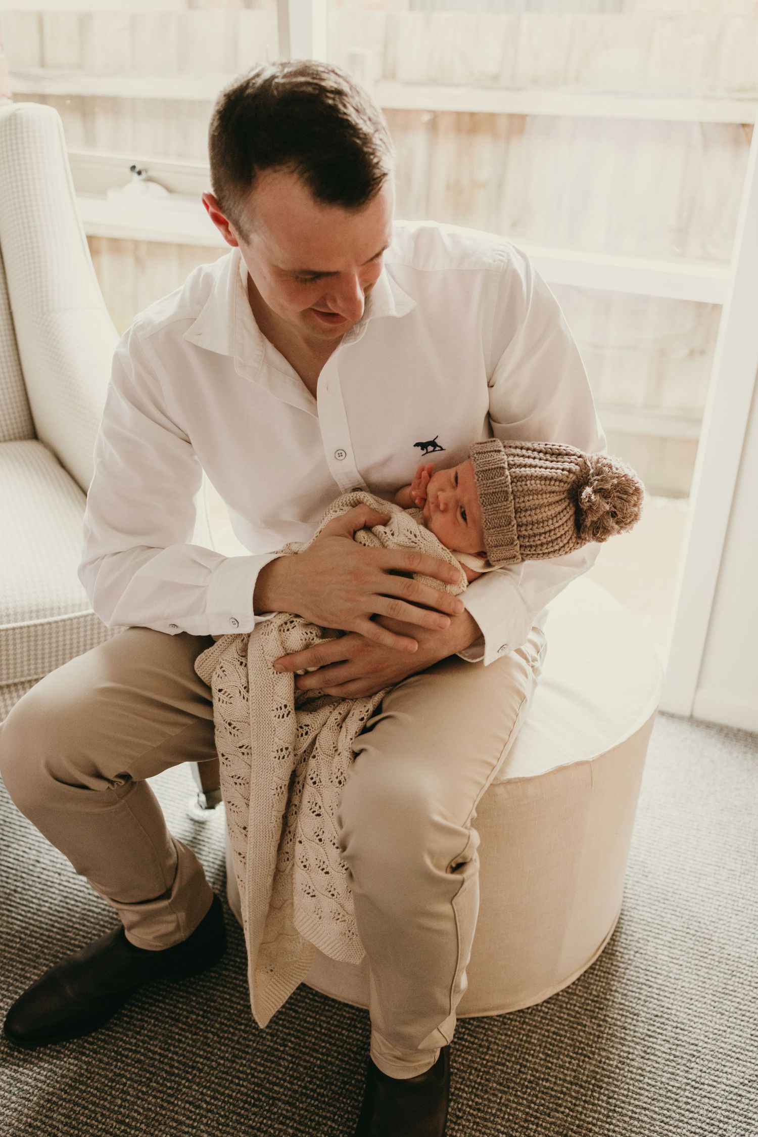 A person in white shirt holding an infant wearing a knit hat while seated in a white chair.