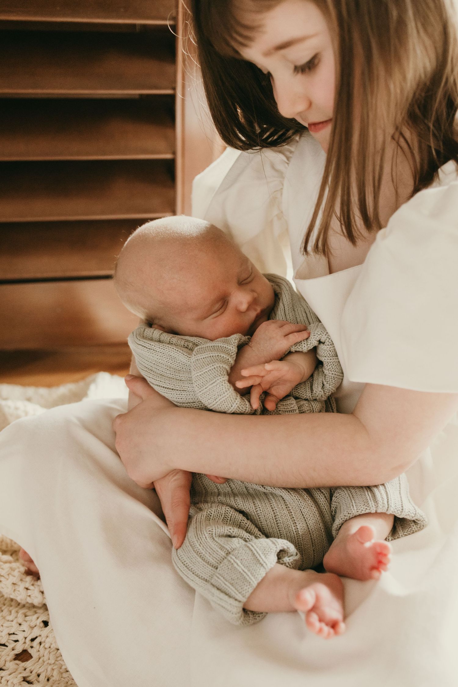 Sleeping newborn in gray knit clothing nestled against caregiver in white dress by window.