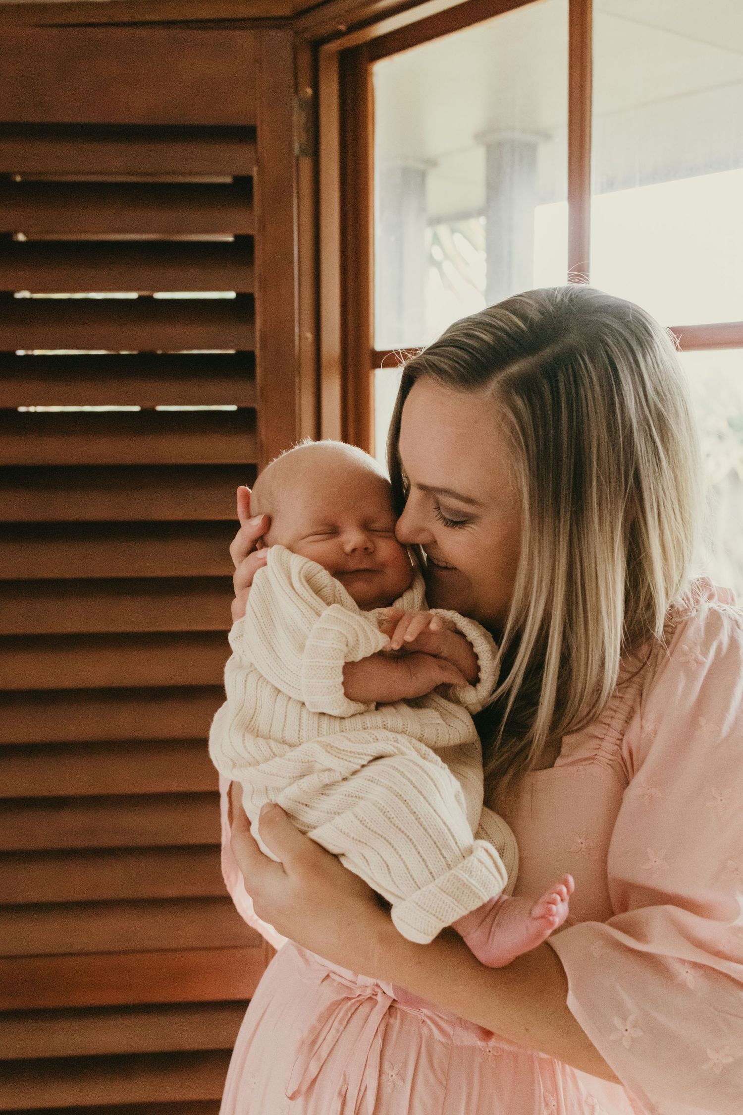A tender moment between a newborn baby in a white knit outfit and caregiver by a window.