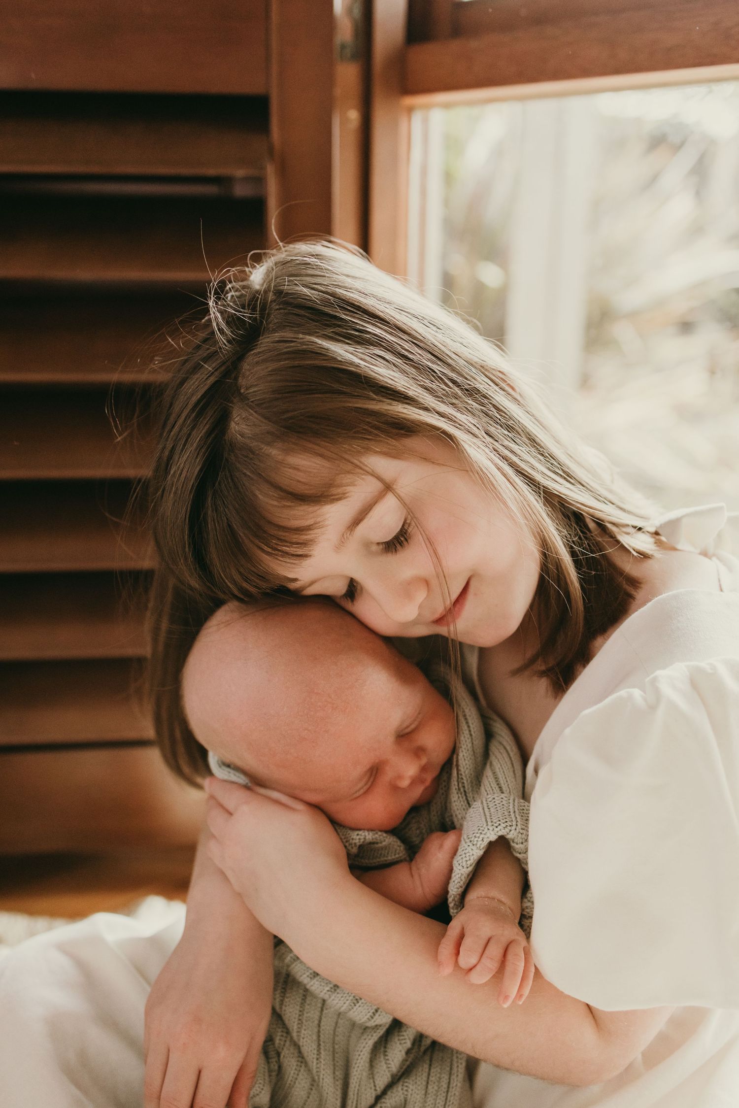 A tender moment captured as a girl in white embraces a sleeping baby near a window with wooden blinds.