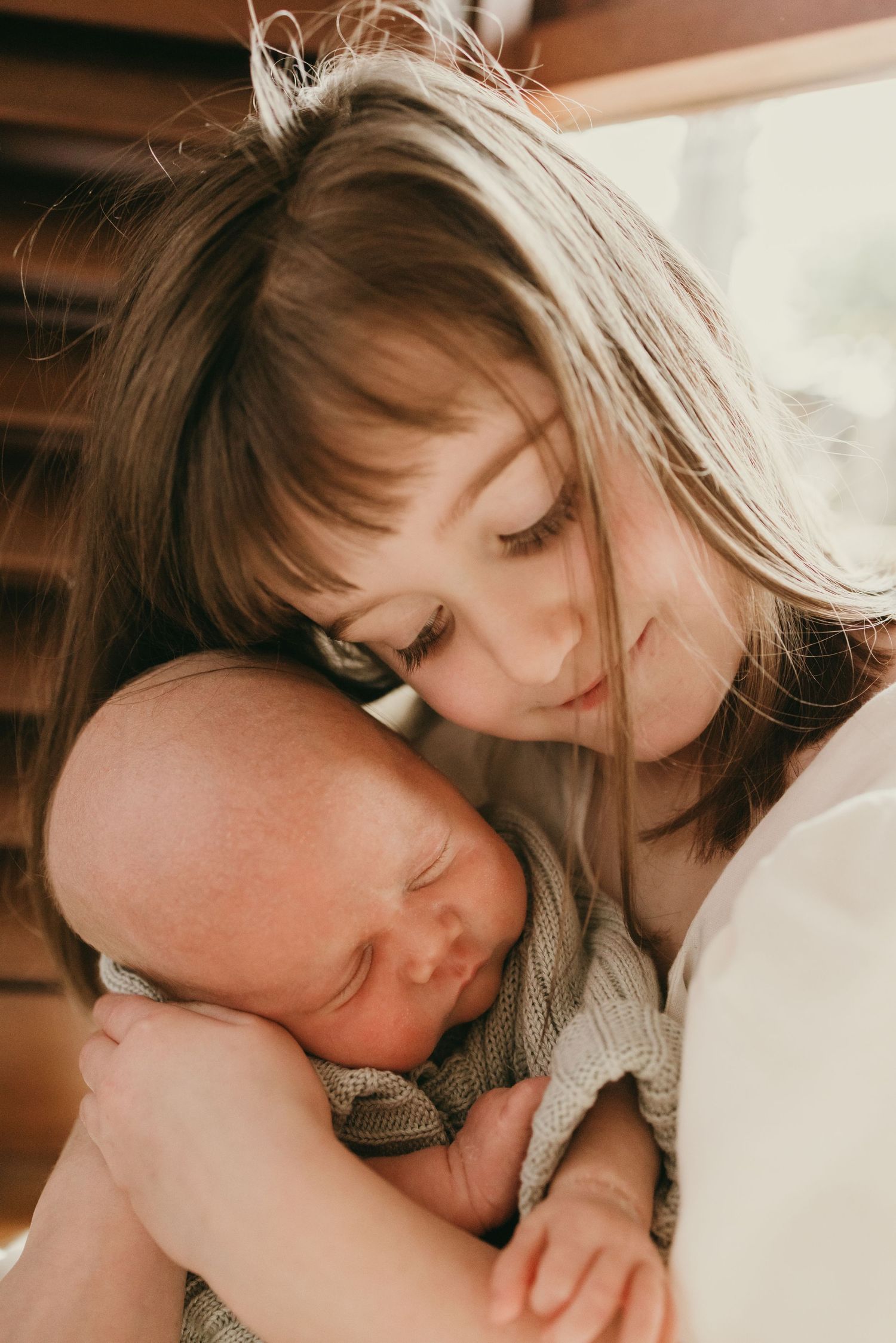 Close-up of a tender embrace between a young child and an infant in natural window light.