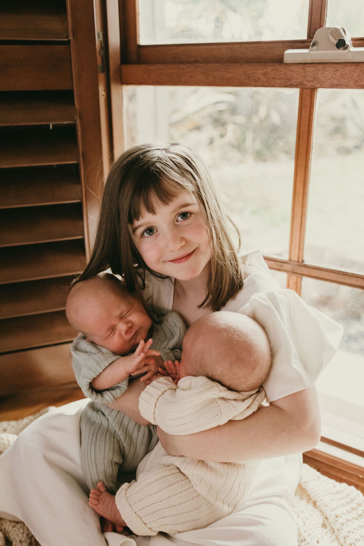 Sweet moment by a window as a child in white holds two sleeping infants.
