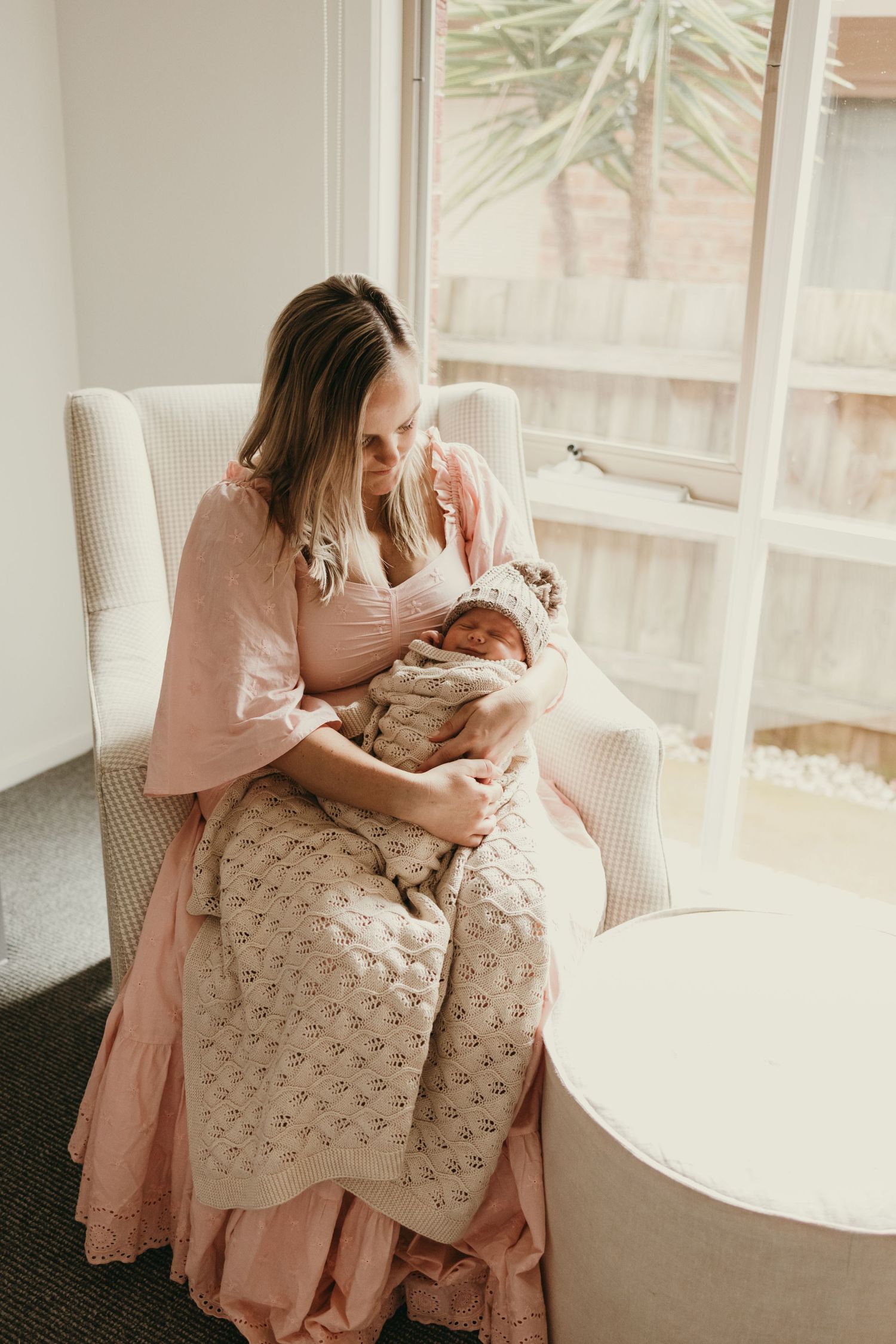 Mother in pink dress sitting in white chair by window holding sleeping newborn.