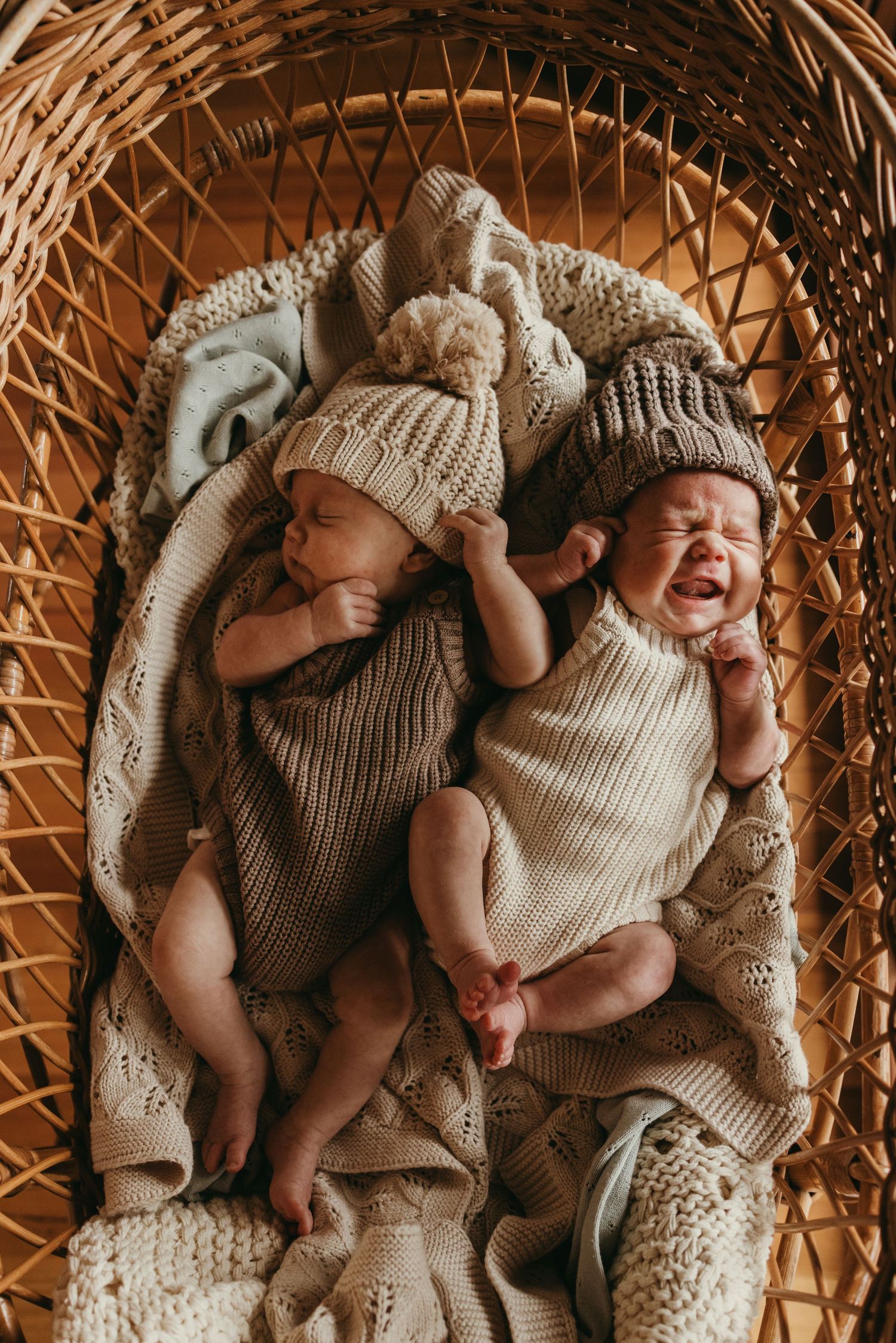 Two newborn babies in knit outfits sleeping peacefully in wicker bassinet.