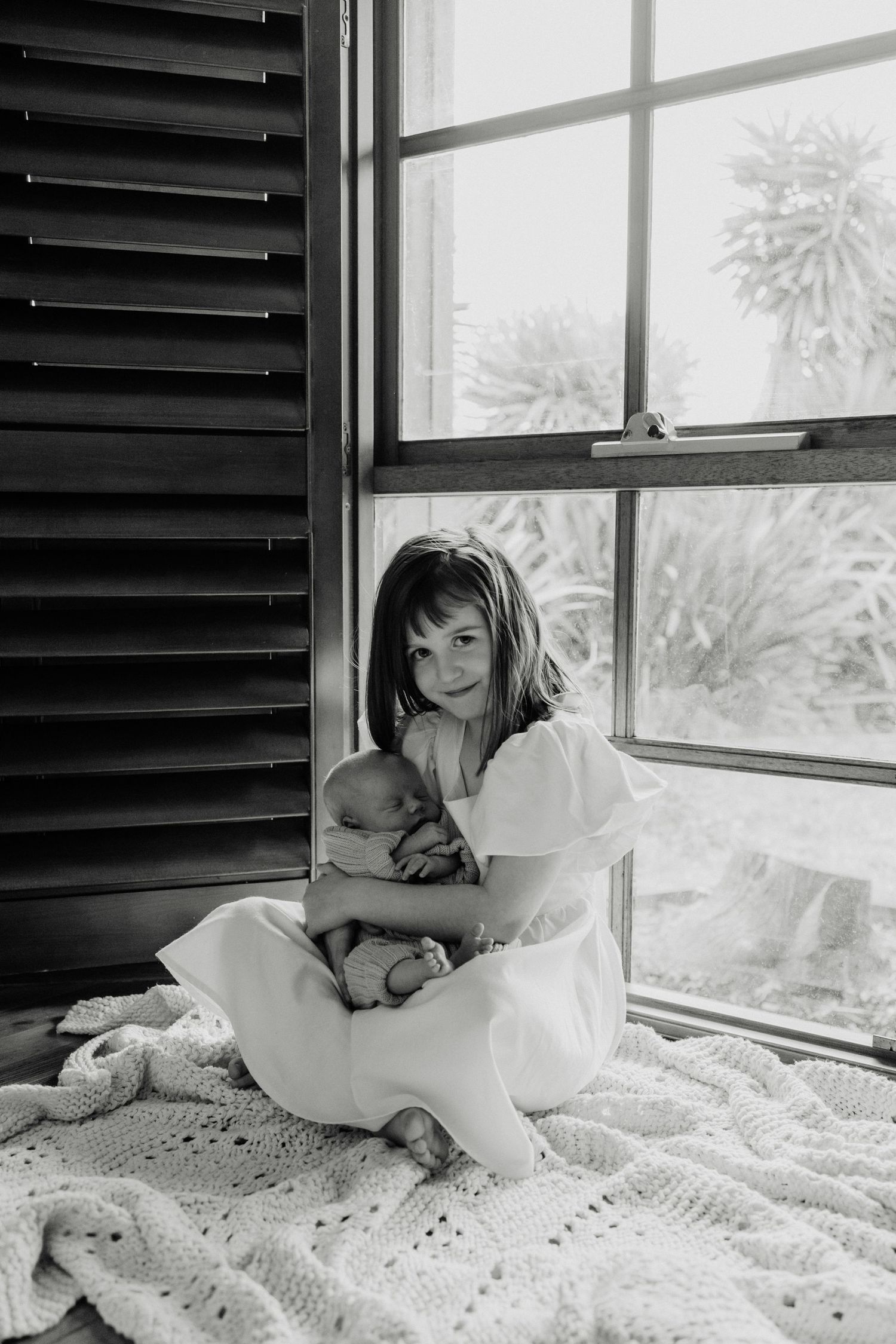 Black and white portrait by window showing gentle embrace with sleeping newborn.