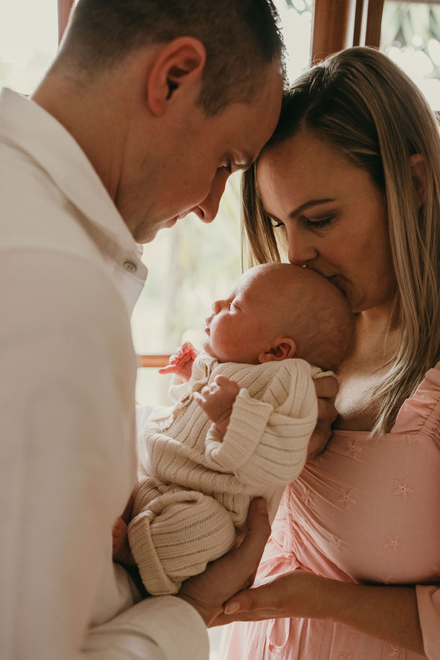 Intimate family moment with parents cradling their newborn baby in soft window light.