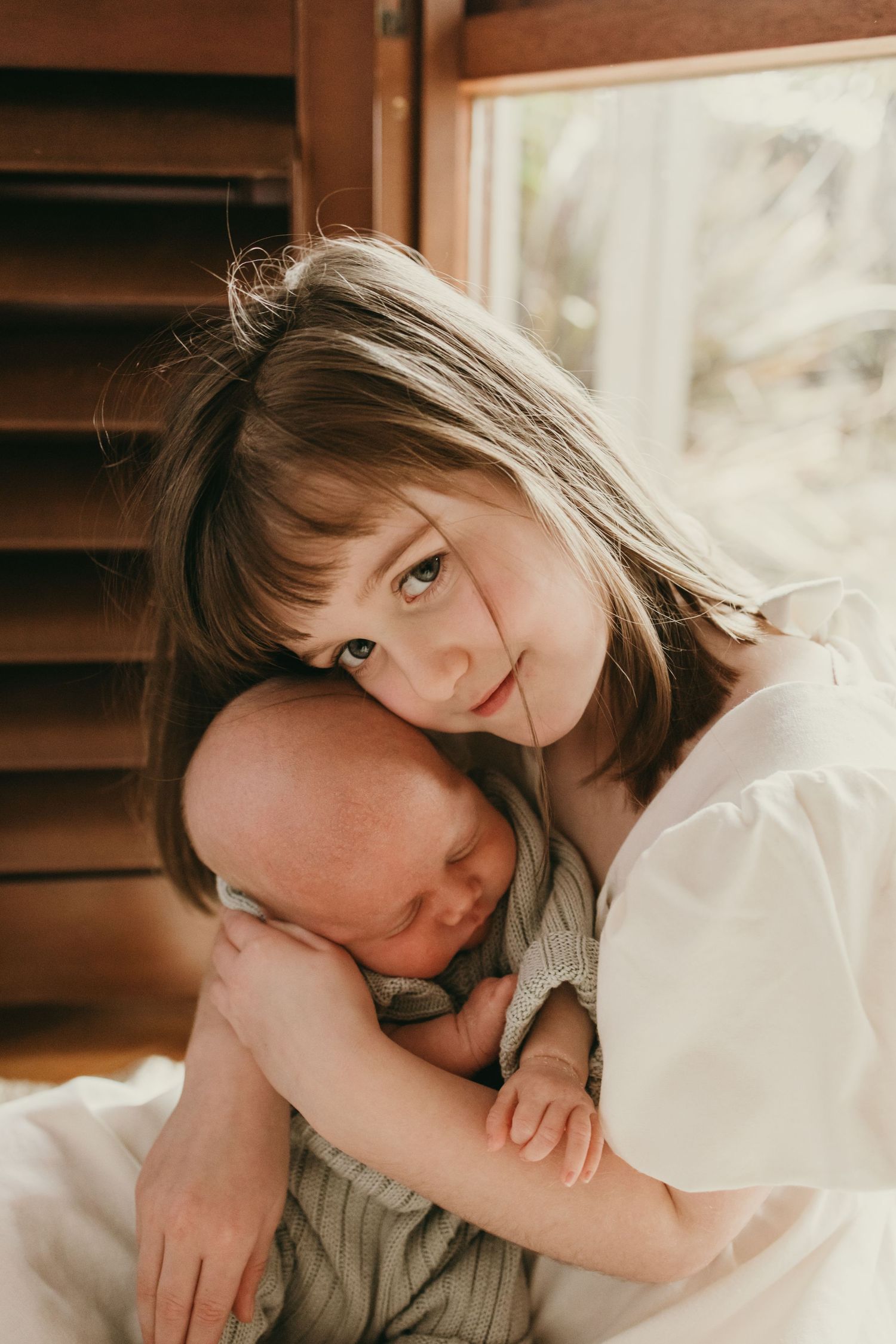 Warm-toned portrait showing peaceful moment with sleeping infant by window with wooden shutters.