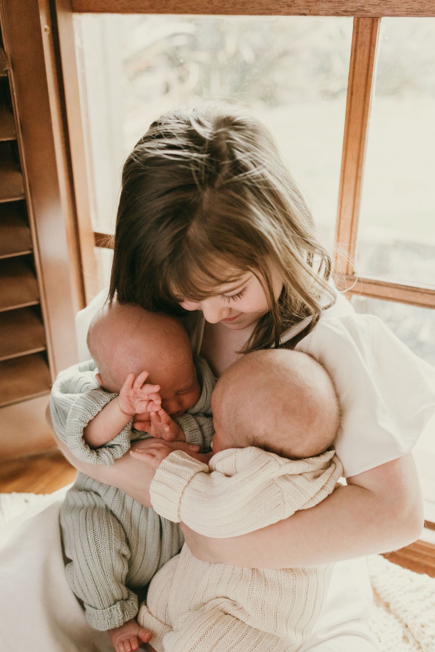 Heartwarming indoor portrait showing a child cradling a sleeping baby by a window.
