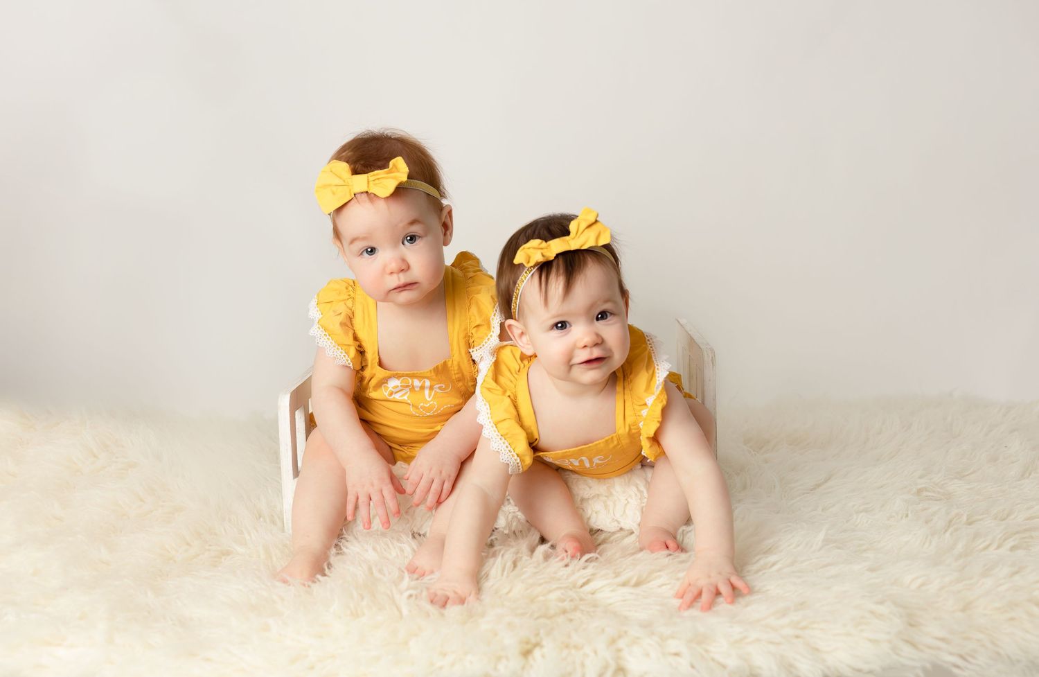 Two infants wearing coordinated yellow outfits and headbands posed on white fur.
