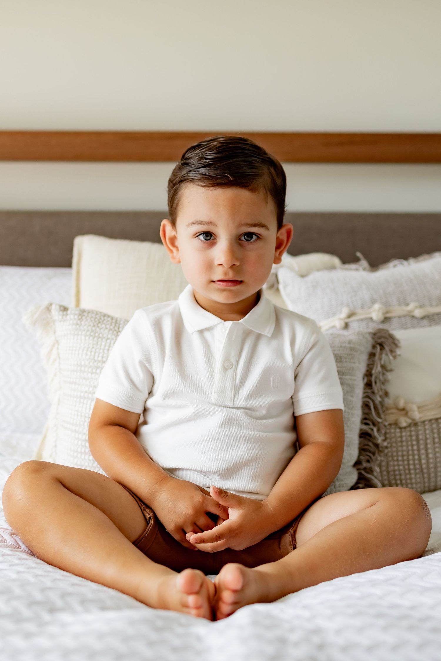 Child in white polo shirt sitting cross-legged on bed with decorative pillows.