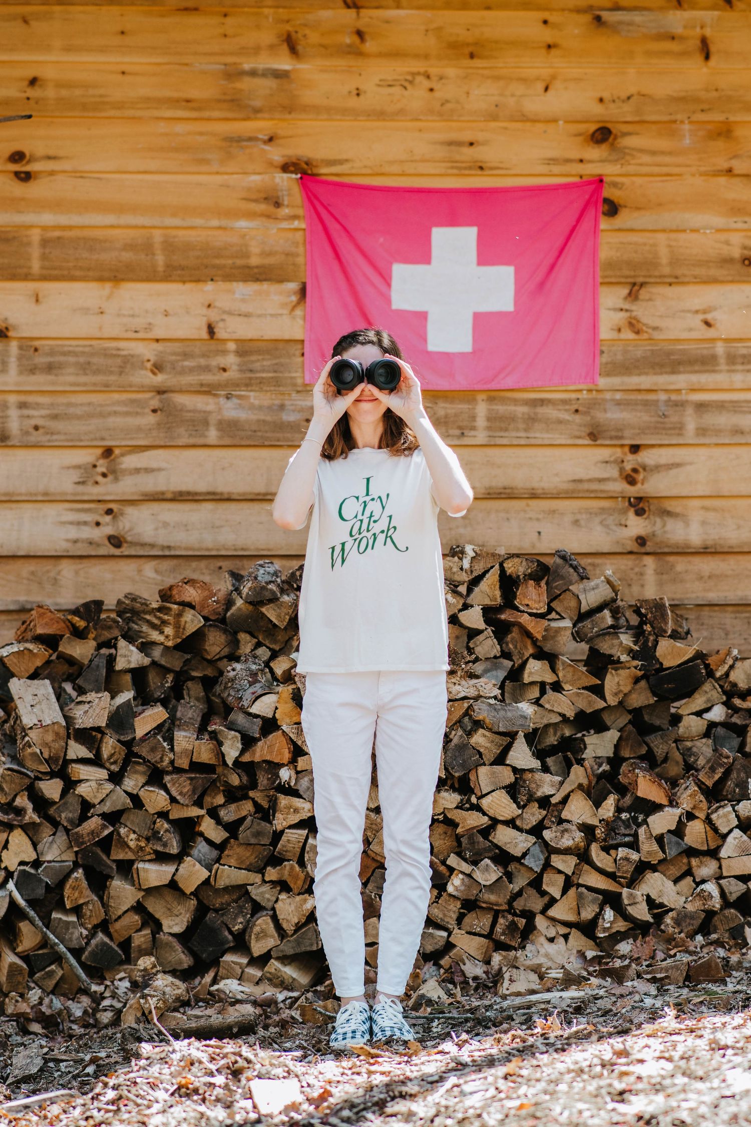 Someone in white winter clothing looks through binoculars against a wooden wall with Swiss flag and stacked firewood in Rhinebeck, New York.