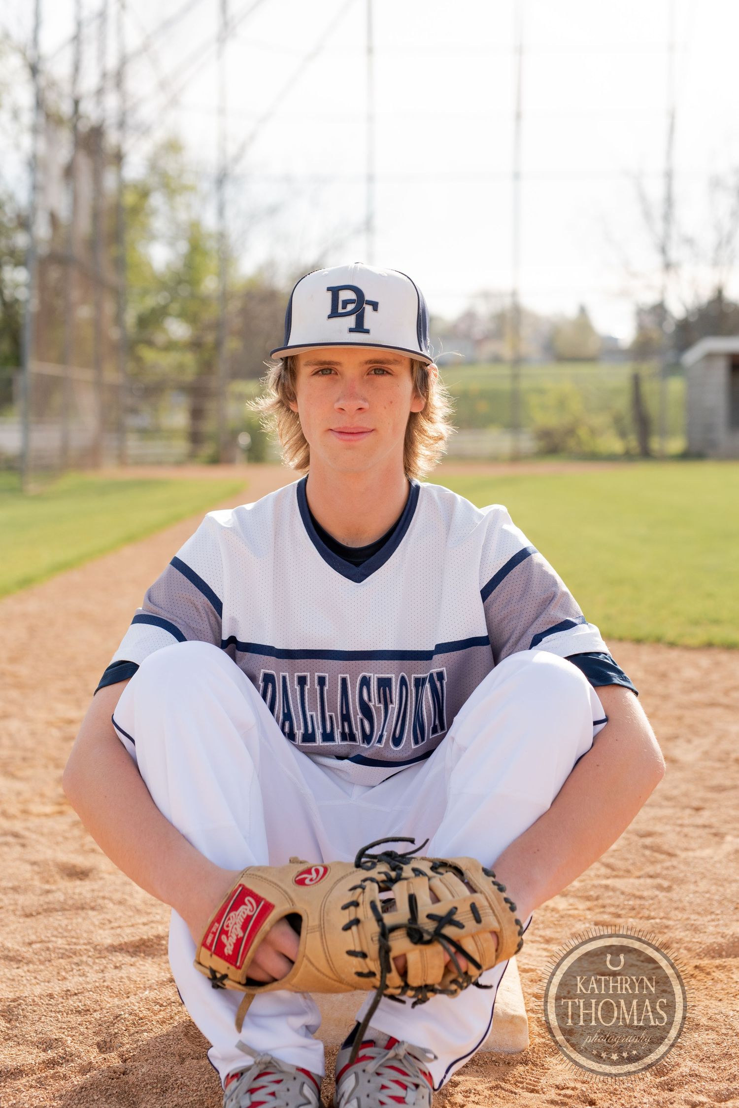 Baseball player in white uniform sits cross-legged with glove on baseball diamond.
