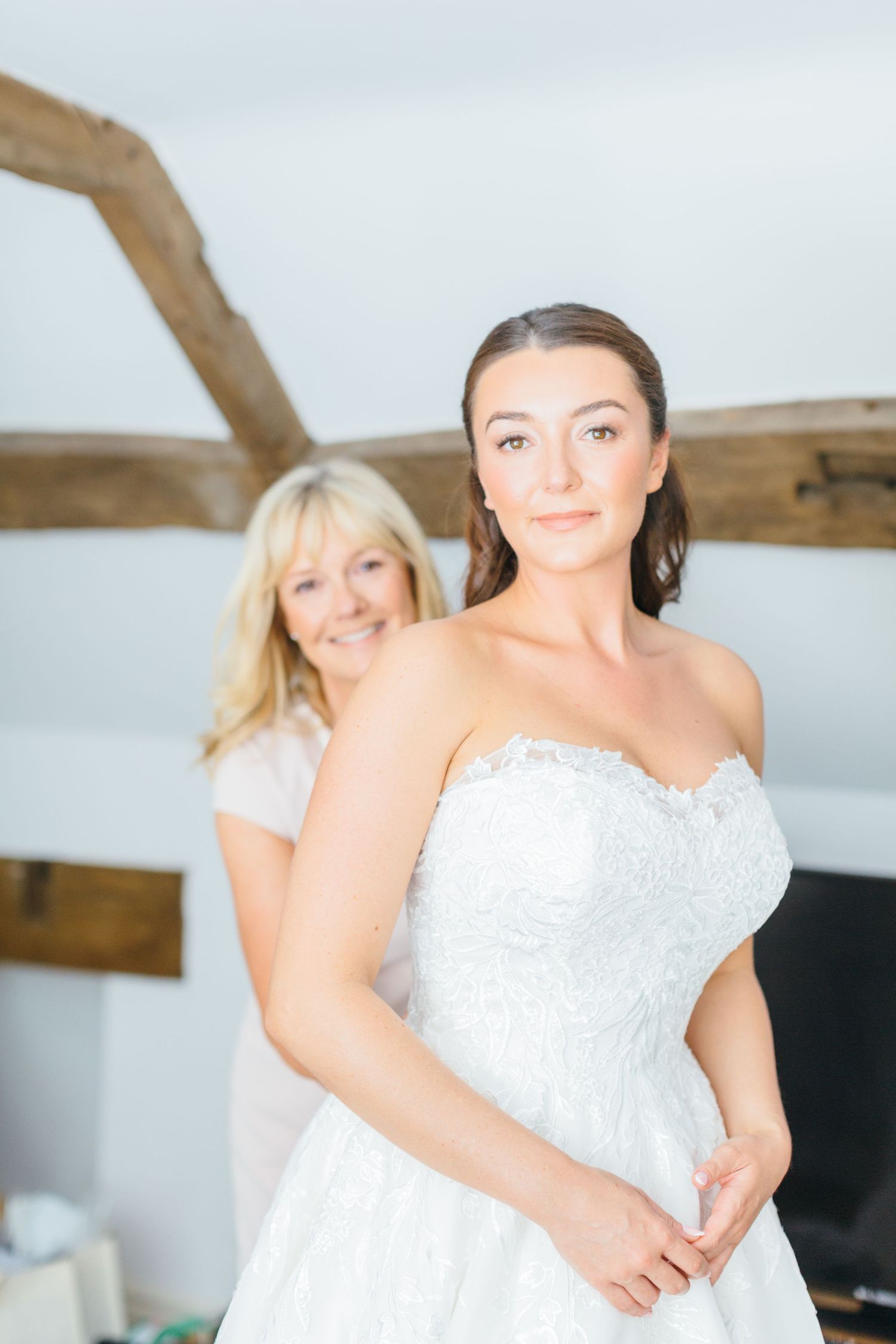 A strapless white wedding dress with lace detailing is showcased in an indoor setting with exposed wooden beams.