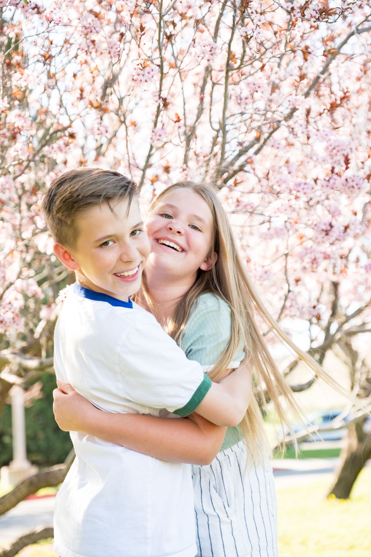 Two children hugging and laughing together under beautiful pink cherry blossom trees on a sunny spring day.
