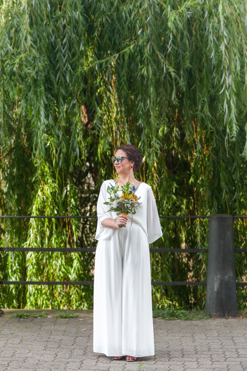 A person in a white dress stands beneath a weeping willow tree holding greenery.