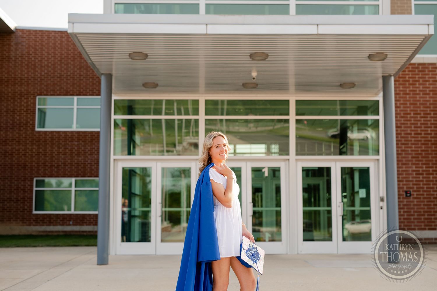 Graduate stands by glass entrance doors wearing blue graduation gown and white dress.