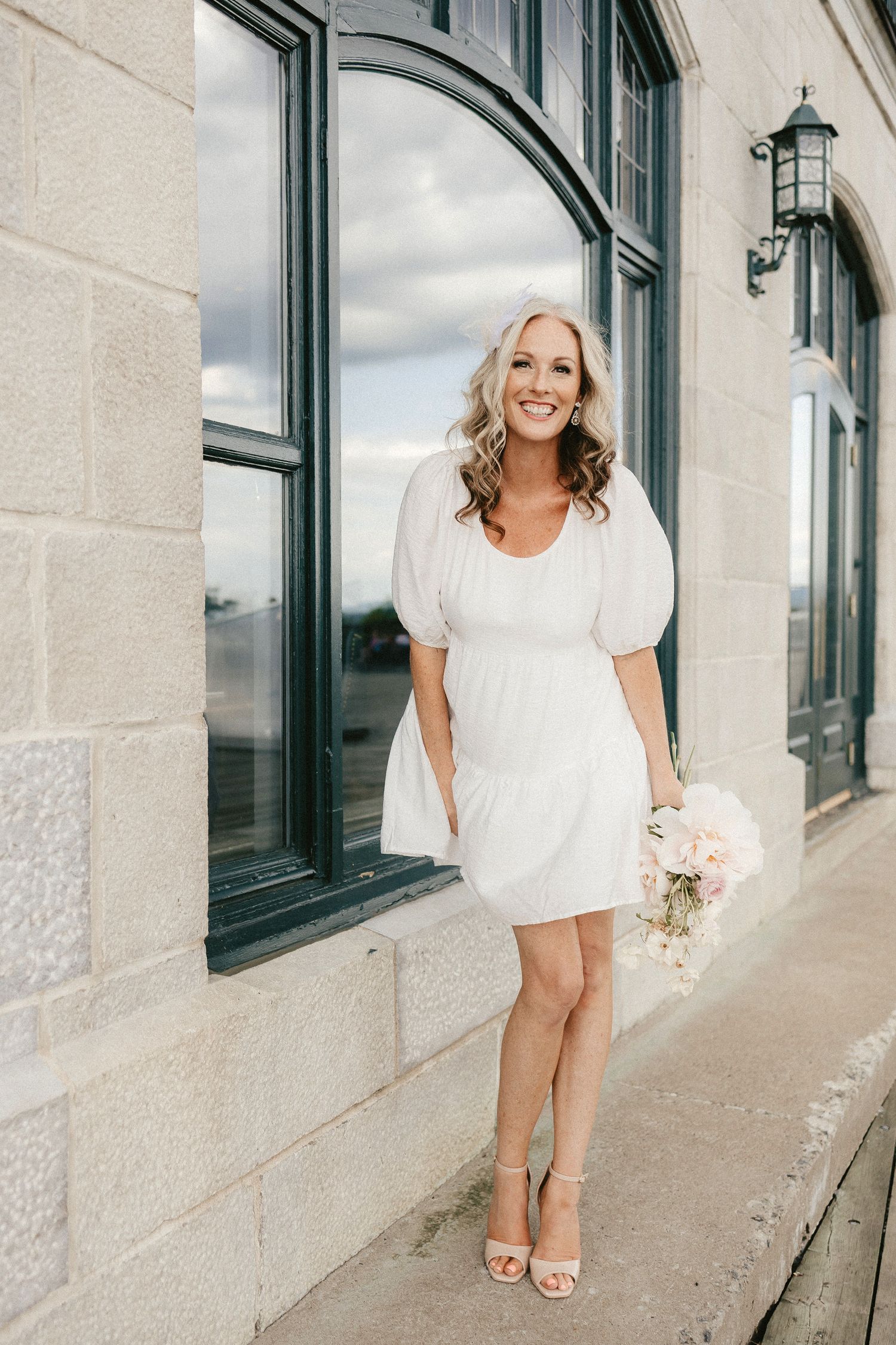 A short white wedding dress styled with puff sleeves and strappy sandals against stone architecture.