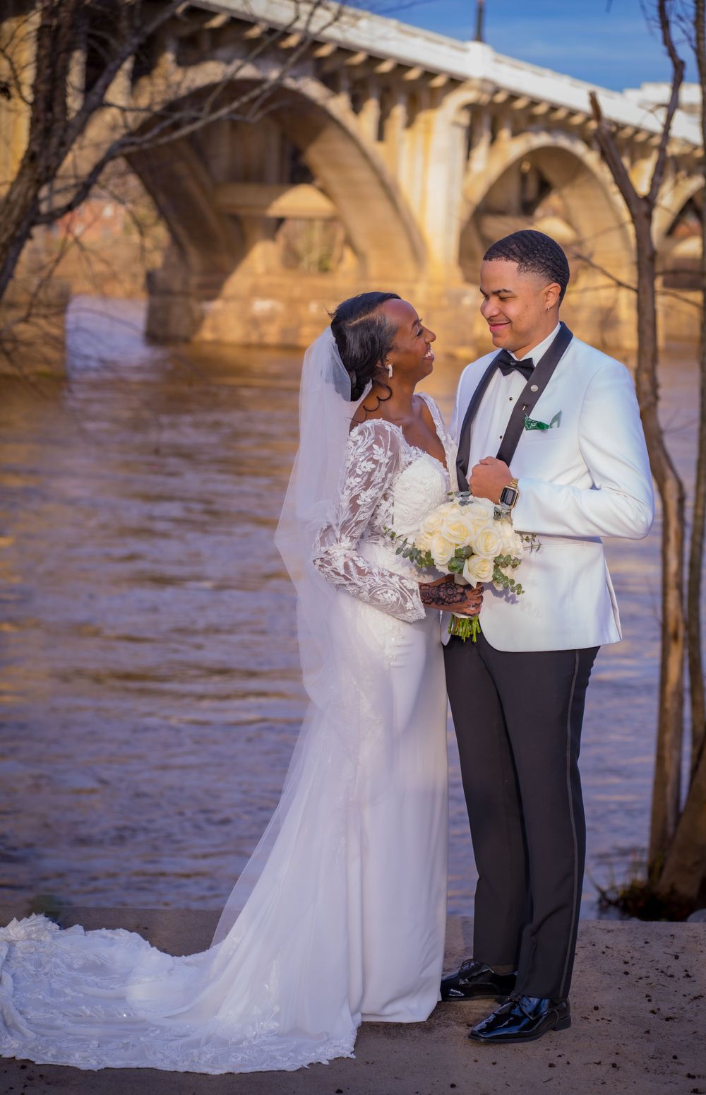A wedding couple poses under an arched bridge with flowing water in the background.