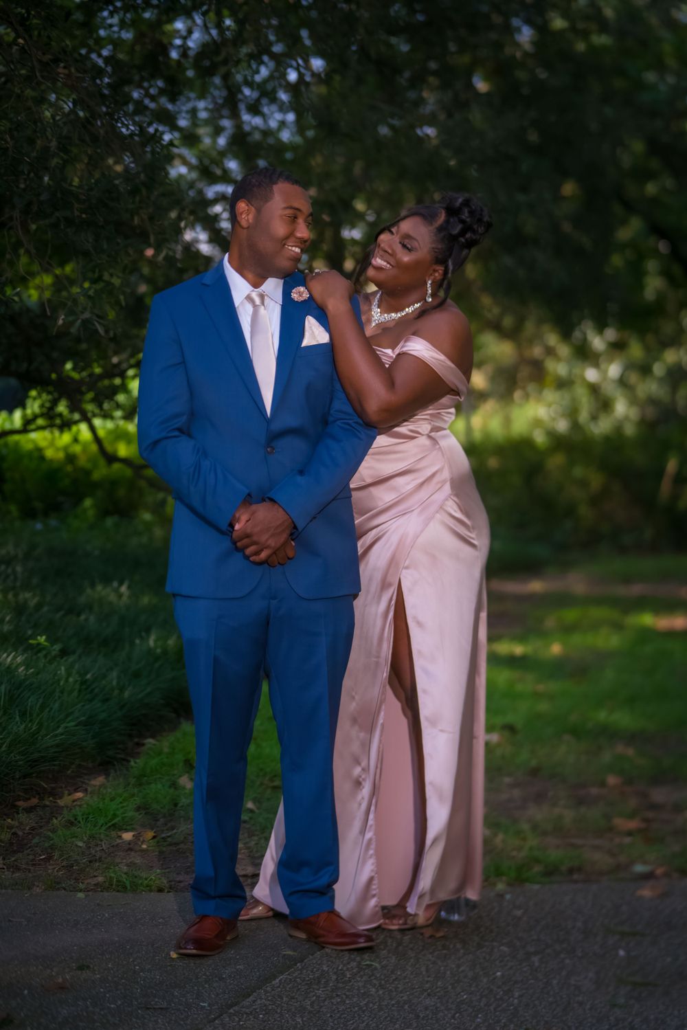 A joyful couple share a candid moment outdoors, with one wearing a blue suit and the other in a pink dress.