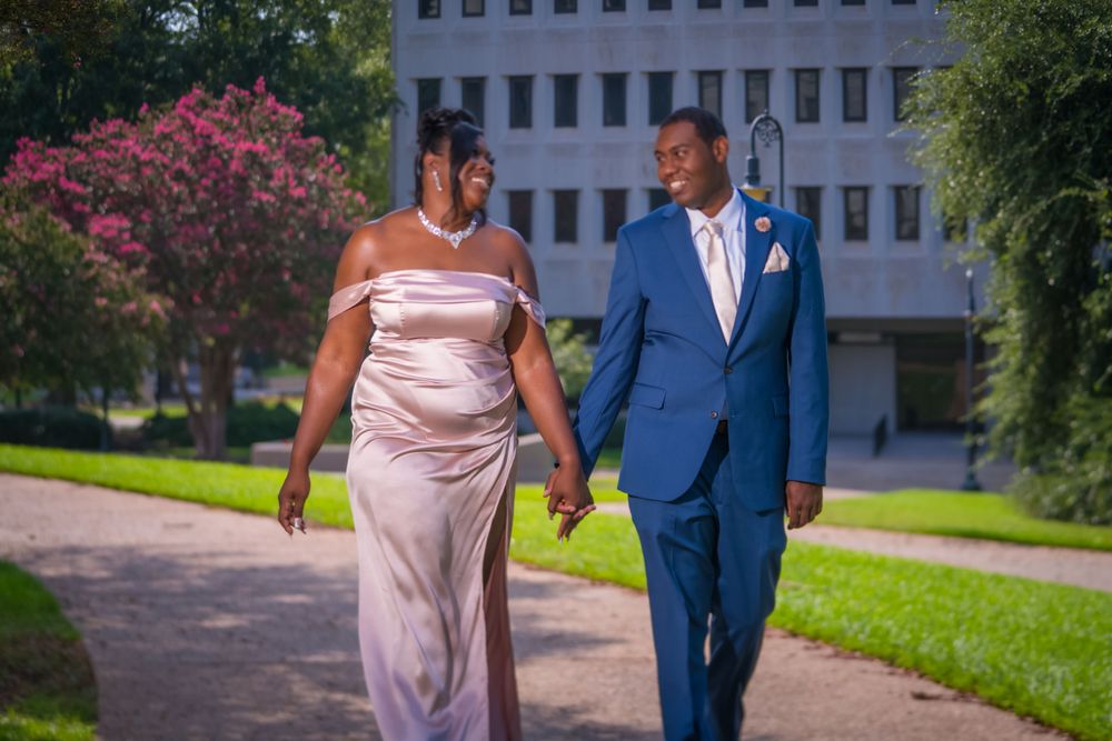 A couple walk together on a garden path, dressed formally in blue suit and pink gown.