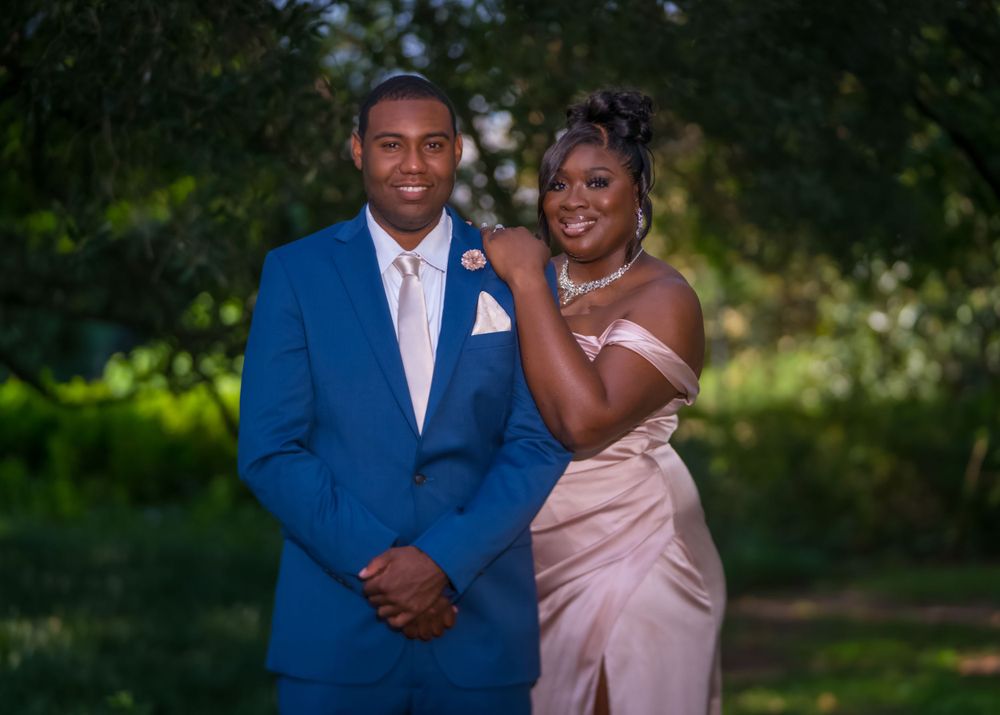 A couple pose together for a formal photo, wearing a blue suit and pink gown in an outdoor garden setting.