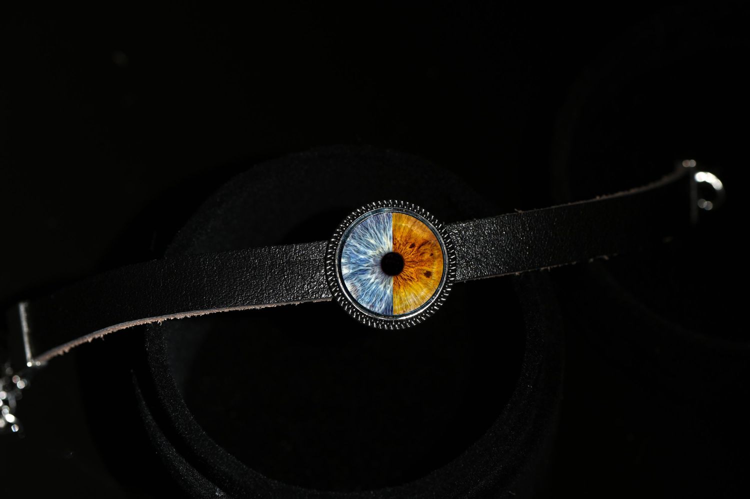 Black leather bracelet with blue and amber iris design pendant on dark background.