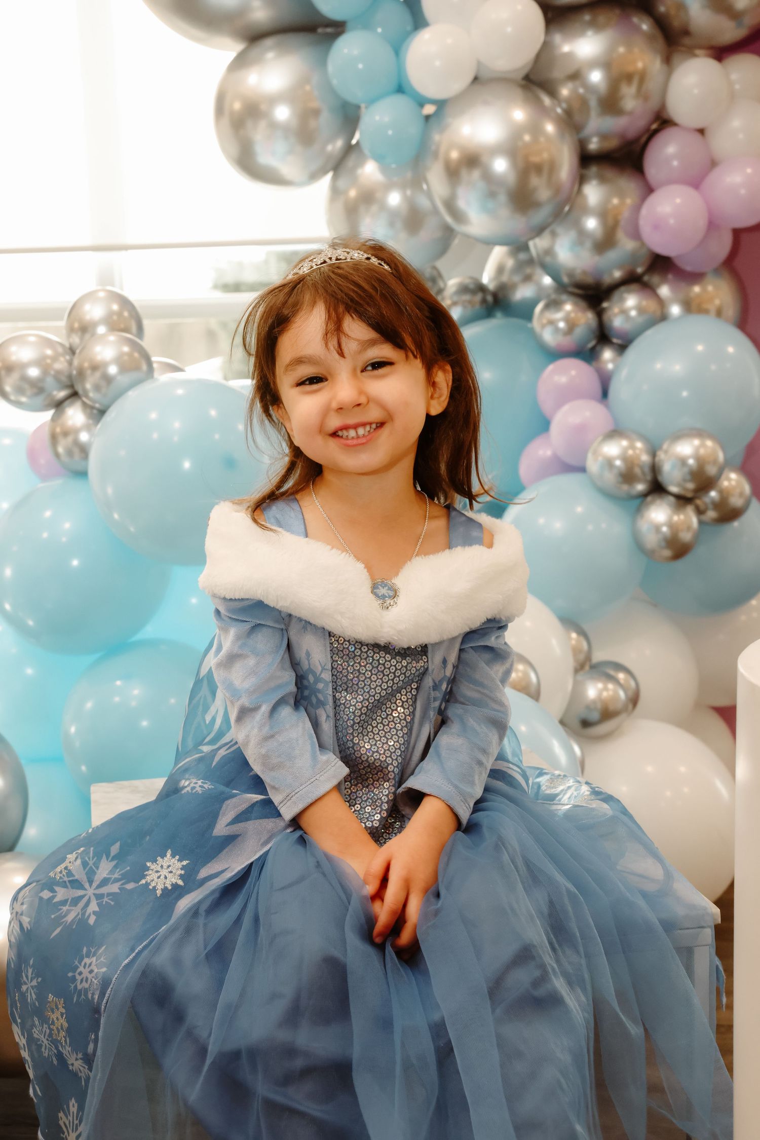 A child wearing a blue princess dress poses in front of silver and blue balloon decorations.