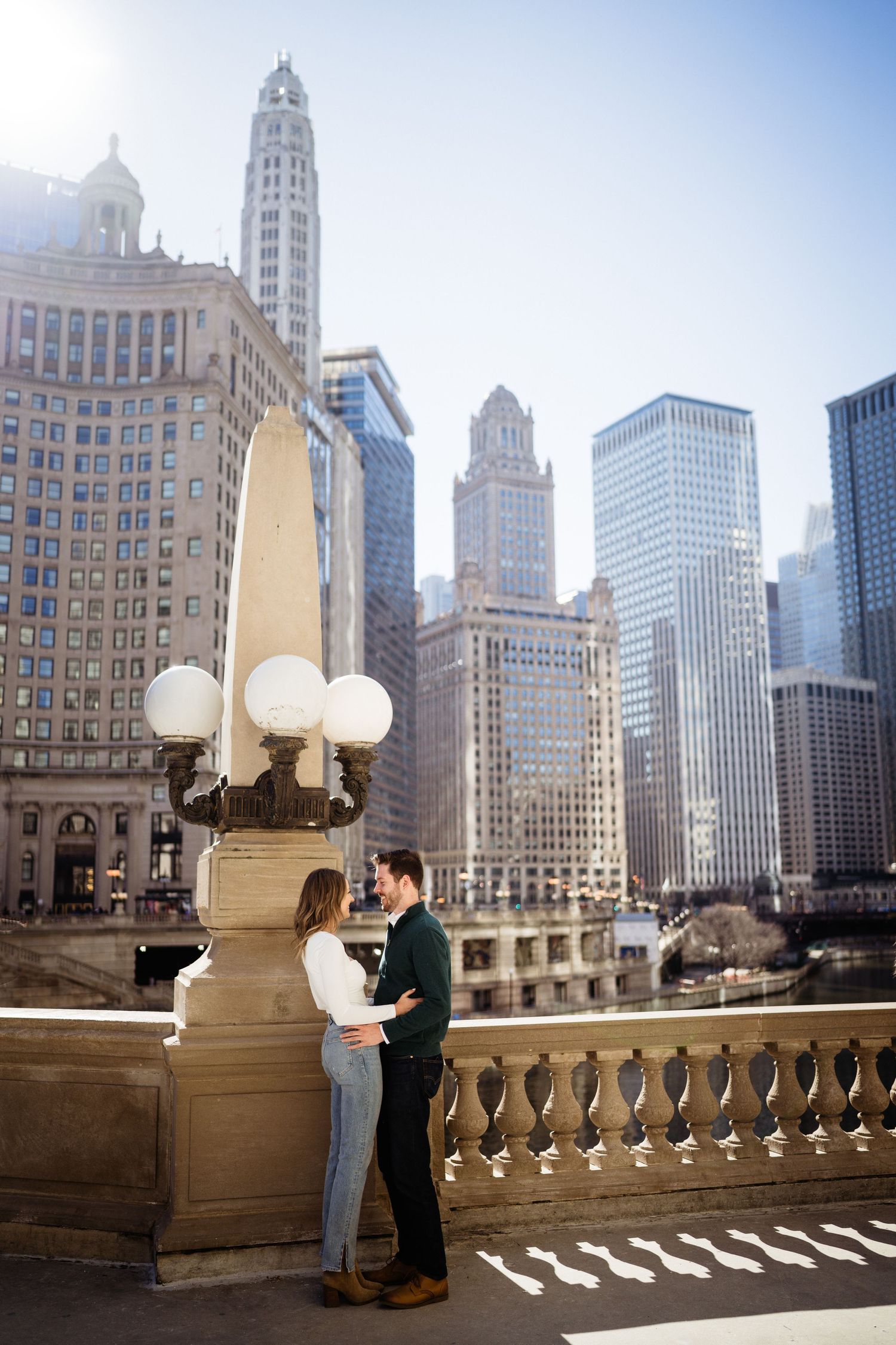 Couple standing close together on bridge with Chicago architecture and vintage lamp posts in background.