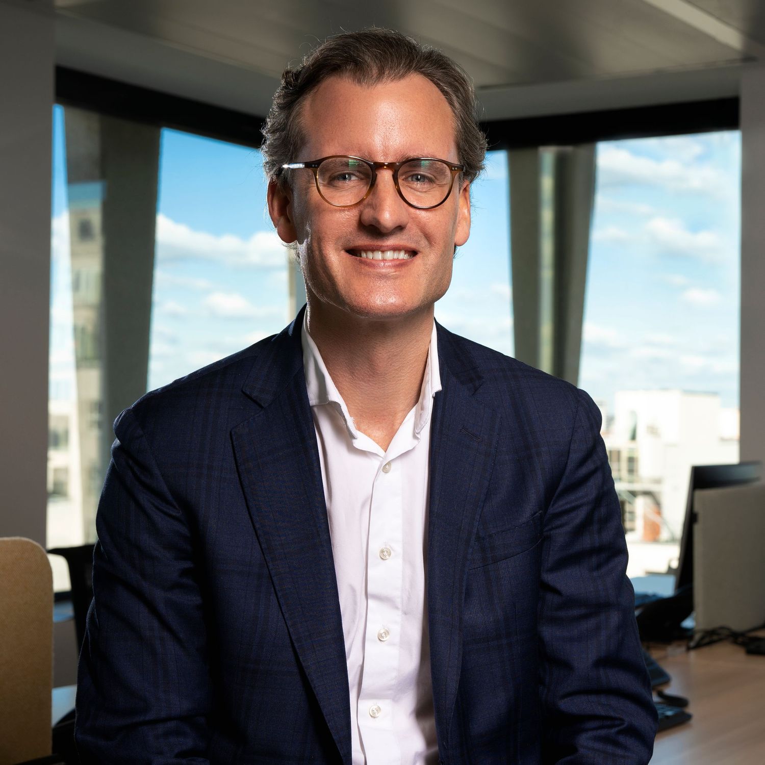 Professional portrait of a person in business attire seated in an office with city views.