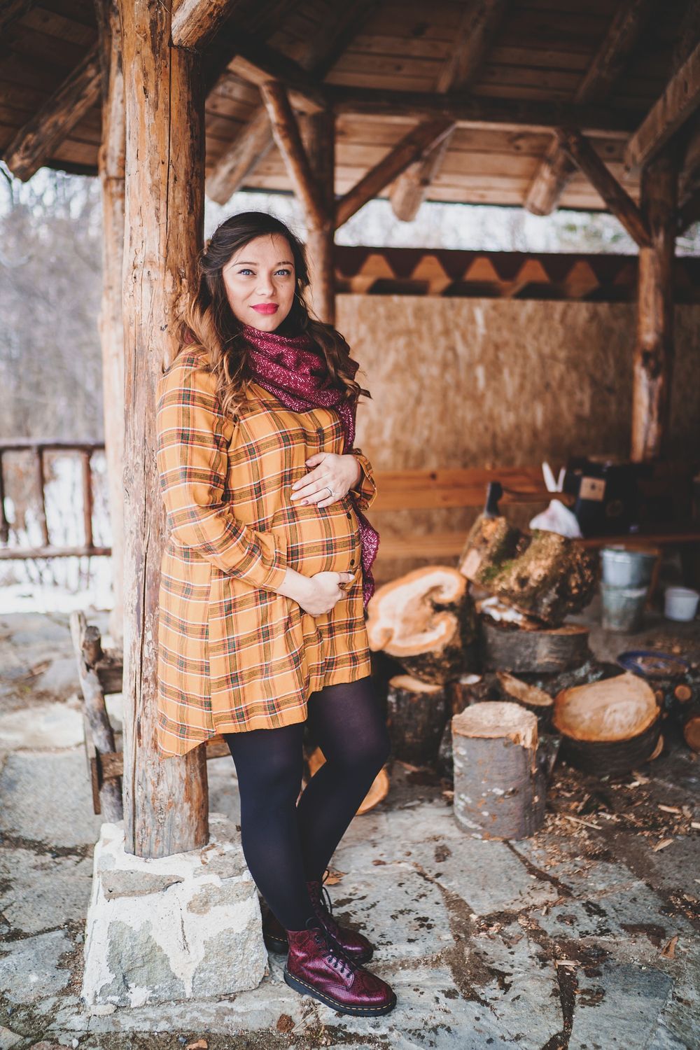 A winter maternity photoshoot featuring a plaid dress and burgundy scarf in a rustic wooden cabin setting.