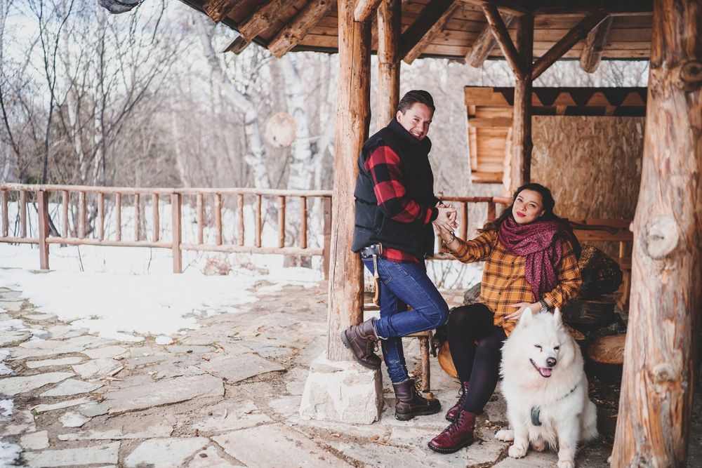 Winter scene of couple sitting in wooden shelter with white dog beside them.