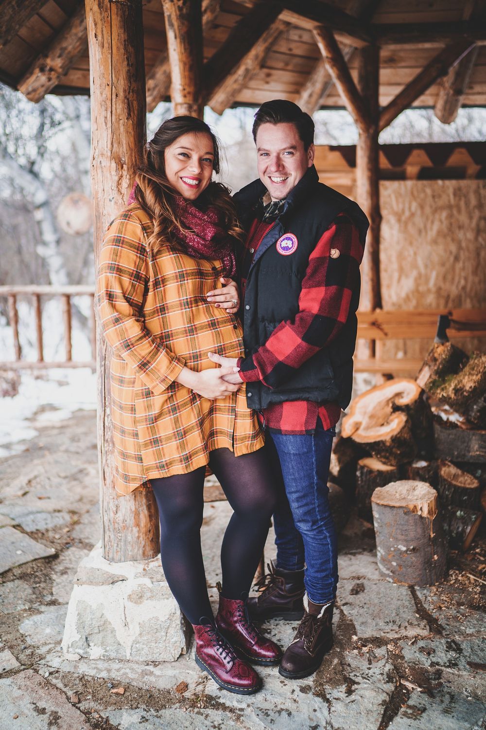 A cozy winter maternity photo with couple wearing plaid and buffalo check patterns on a cabin porch.