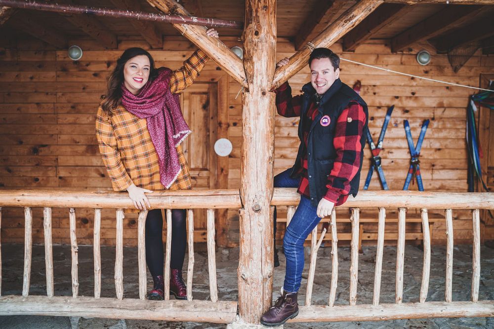 Couple poses playfully on wooden log cabin balcony wearing winter attire.