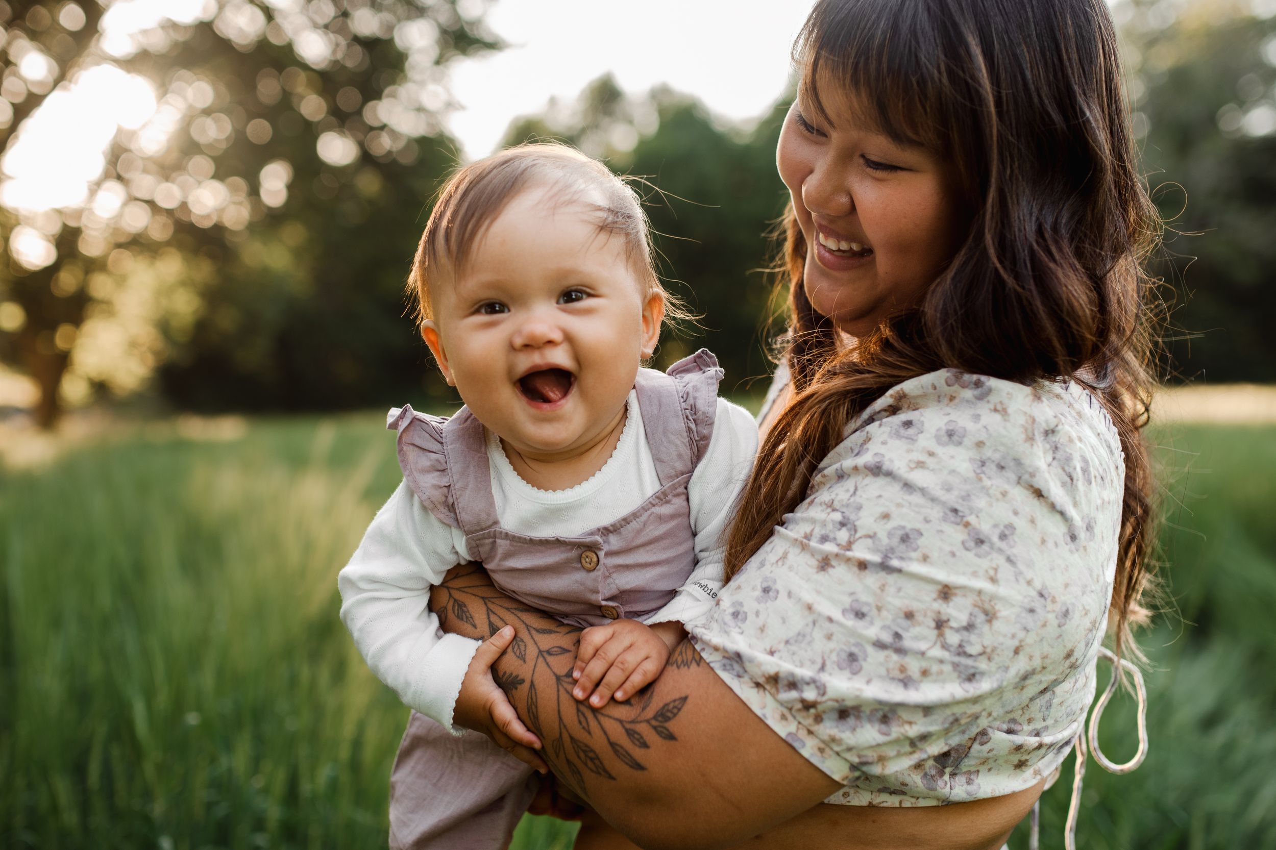 Baby laughing in pink dress while being held outside in a grassy field at sunset.