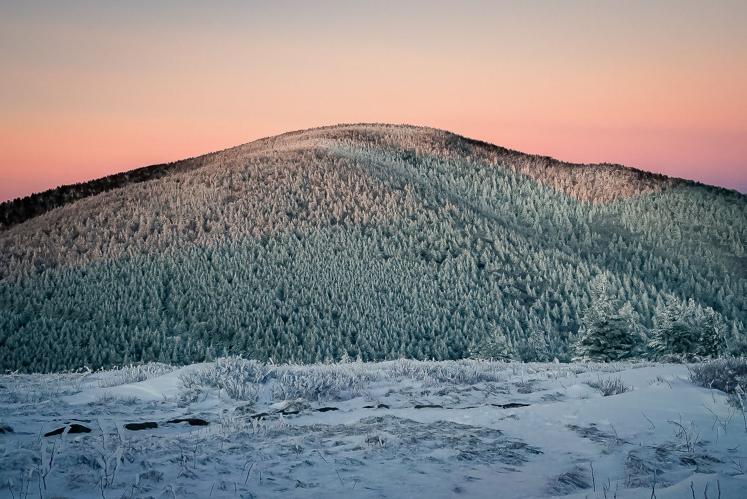 A rosy pink glow haloes a snow dusted mountain in this winter landscape photograph.