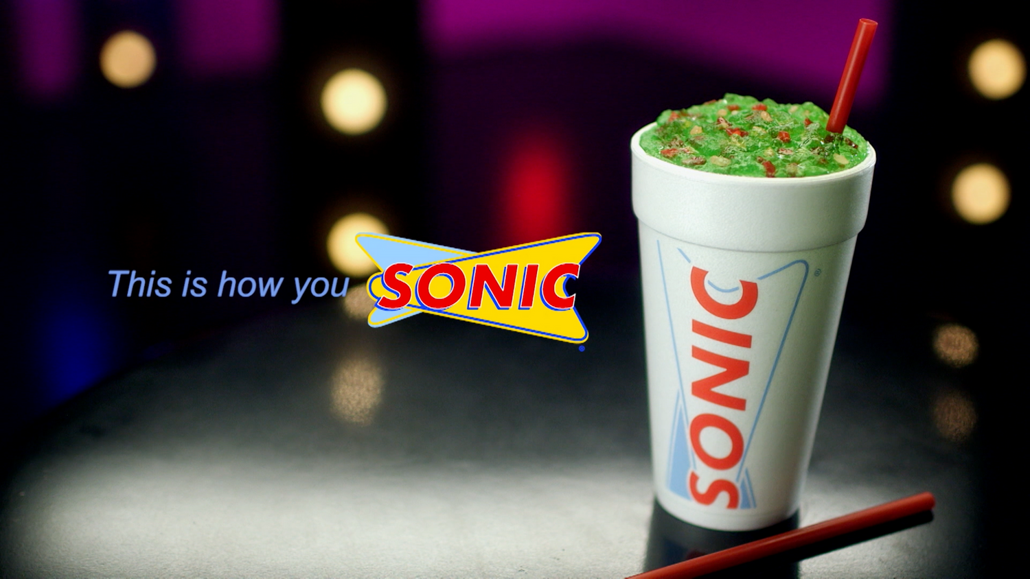 Sonic branded drink cup with green slush beverage against dark background with bokeh lights.