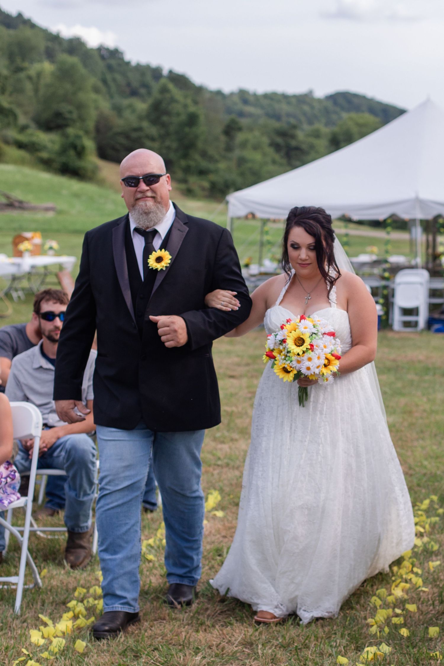 Outdoor wedding ceremony with sunflower decorations and white chairs set up on grassy hillside.