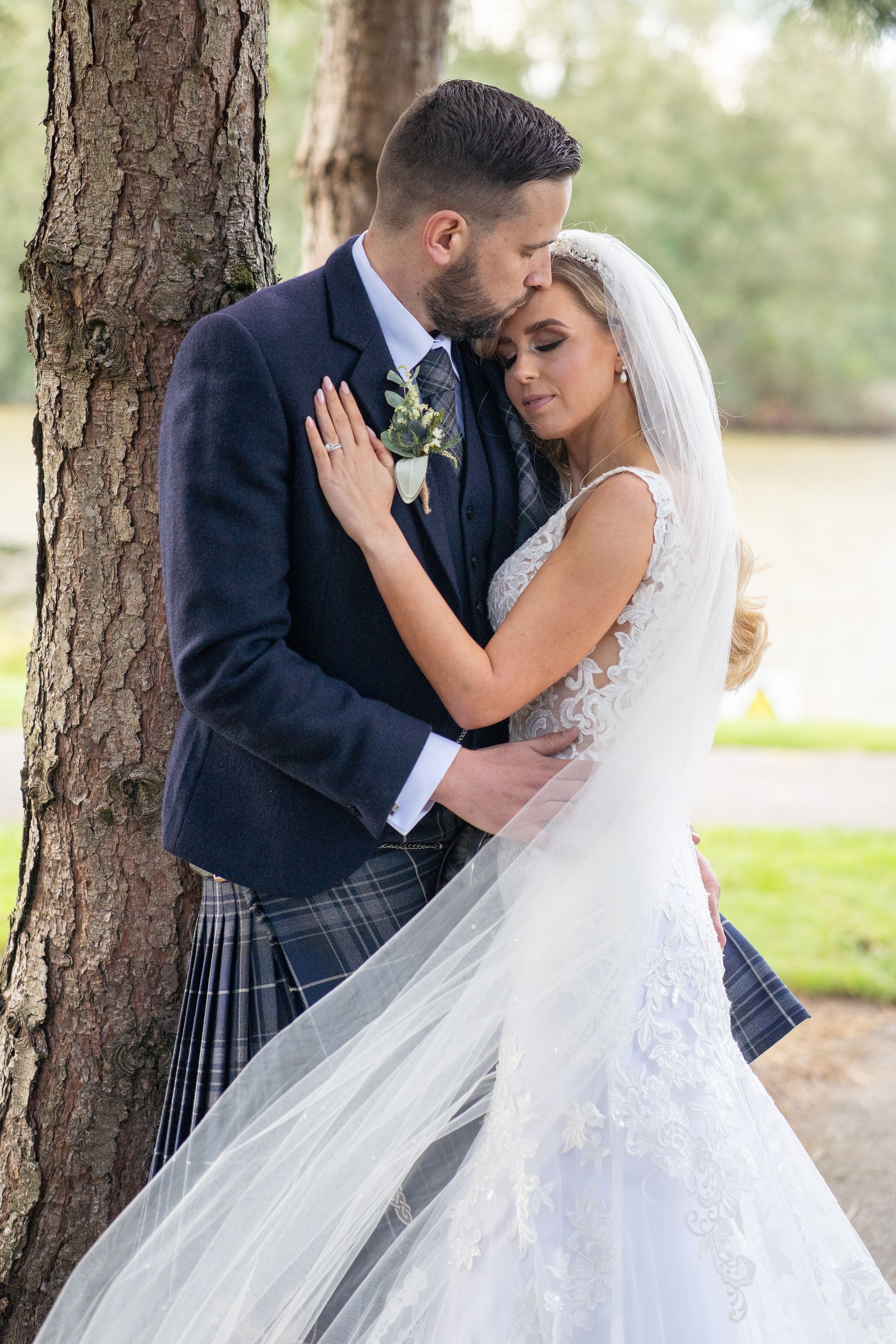 Romantic wedding portrait of couple embracing by tree with bride in lace gown and groom in traditional kilt At Mount Wolseley Hotel