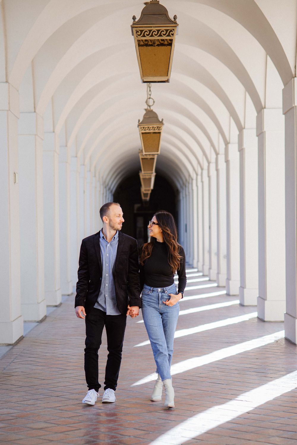 A couple walks through an elegant arched colonnade with dramatic lighting and shadows.