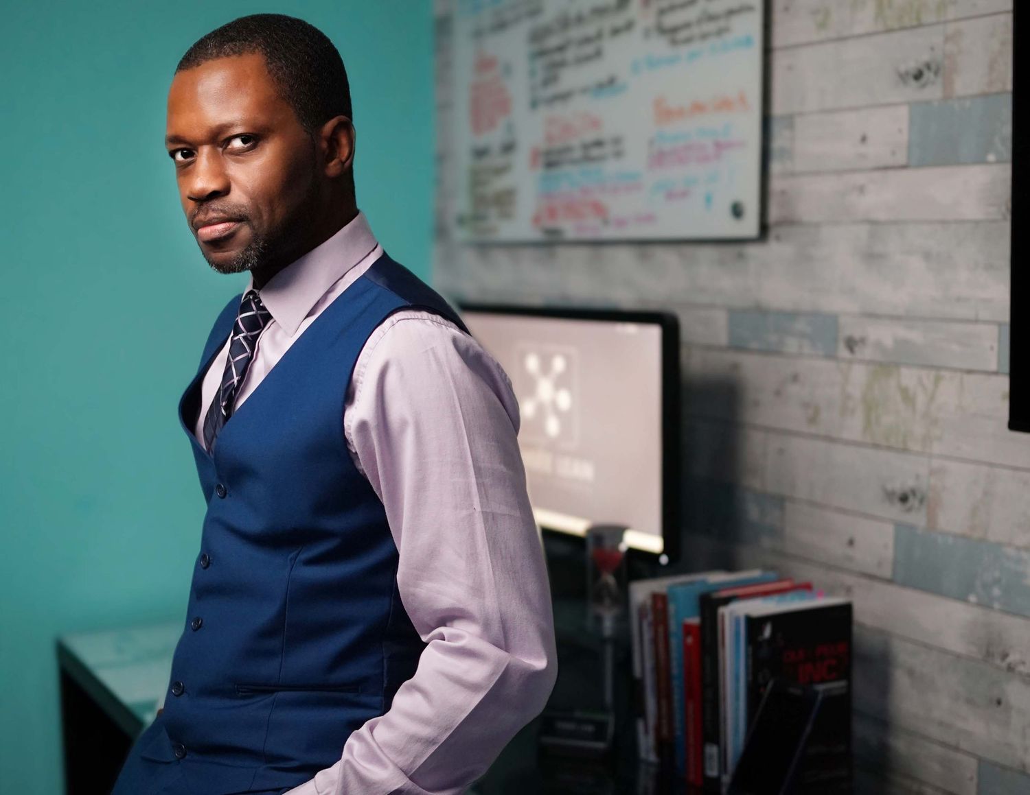 Stylish man in blue vest and tie leaning confidently against a teal wall in modern office.