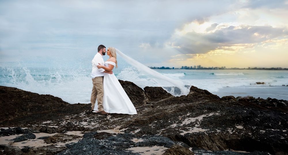 Wedding couple embraces on rocky coastline as veil flows dramatically in ocean breeze.
