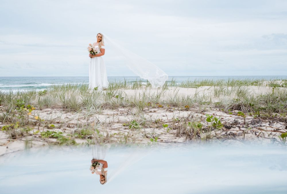 Artistic beach wedding photo with reflection in water against coastal dunes.