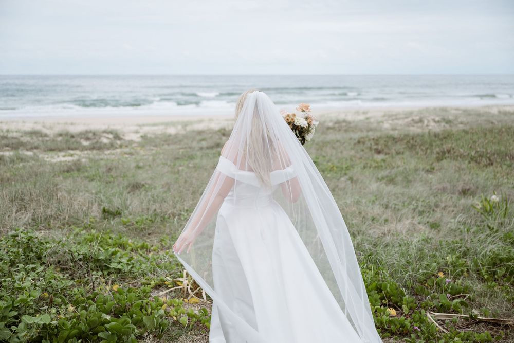 Back view of bride in flowing veil overlooking coastal landscape with bouquet.