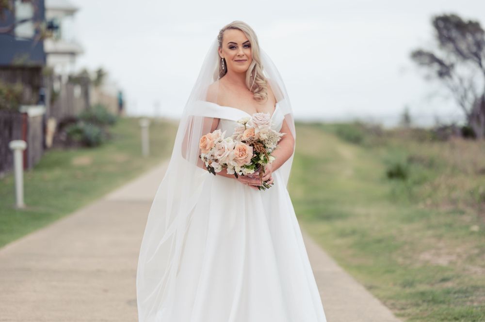Elegant bride walks along coastal path wearing off-shoulder gown and holding peach bouquet.