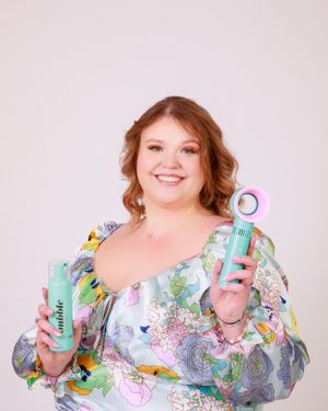 Woman in pastel floral dress holds skincare products while smiling at camera.